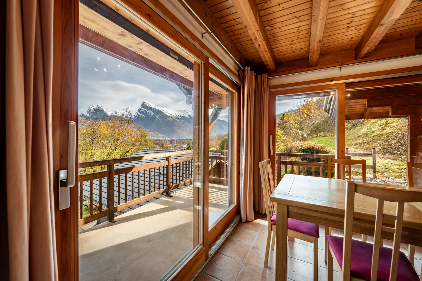 A dining room with a wooden table and chairs, a balcony with a mountain view, and a wooden ceiling.