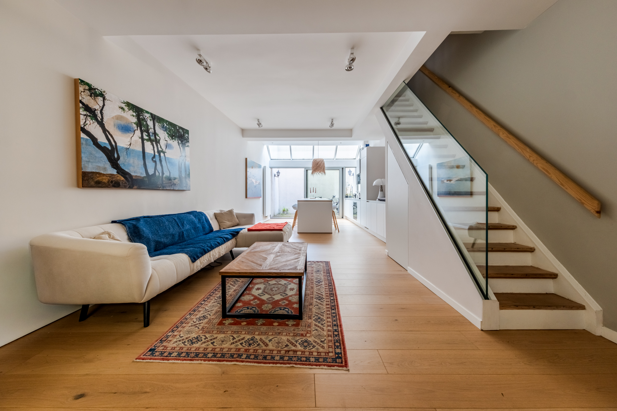 Modern living room with a white sofa, blue throw, patterned rug, wooden coffee table, and stairs with a glass railing. [AI-generated text]