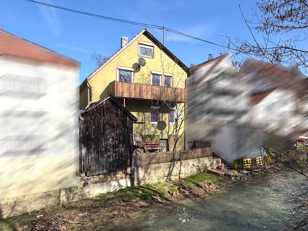 A yellow two-story house with a balcony, a shed, and a small river in front of it.
