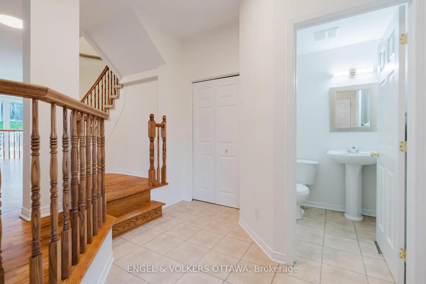Bright foyer with wood stairs, white walls, and beige tile. A half-bath with a pedestal sink is visible through an open door.