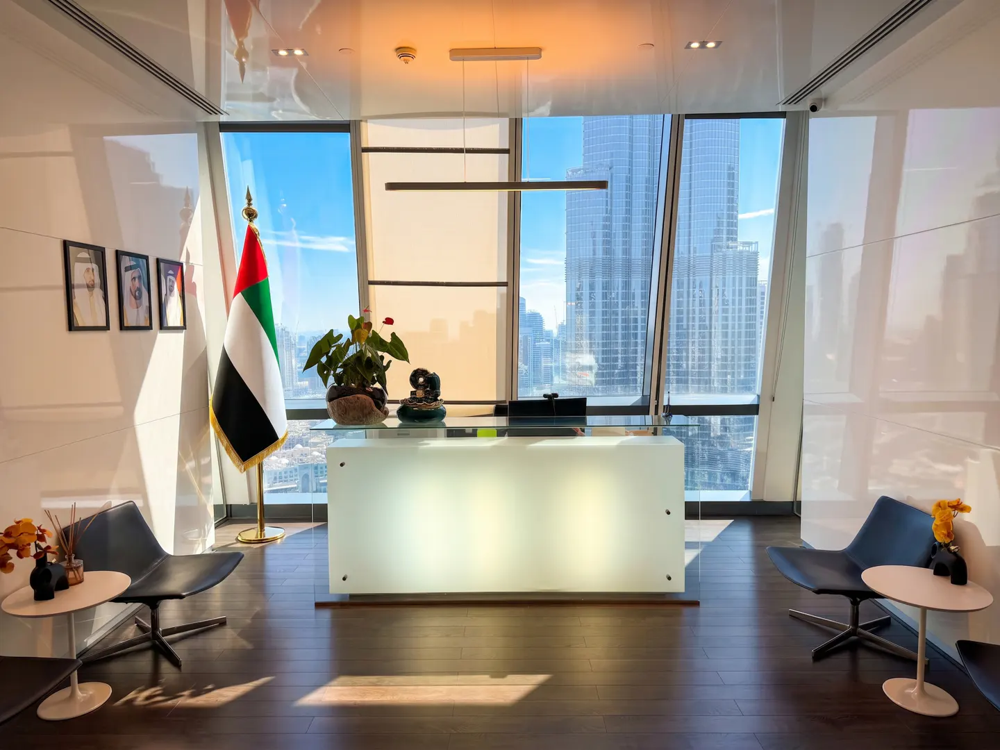 Bright office space with a white reception desk, UAE flag, and city view through large windows. Two chairs and small tables flank the desk.