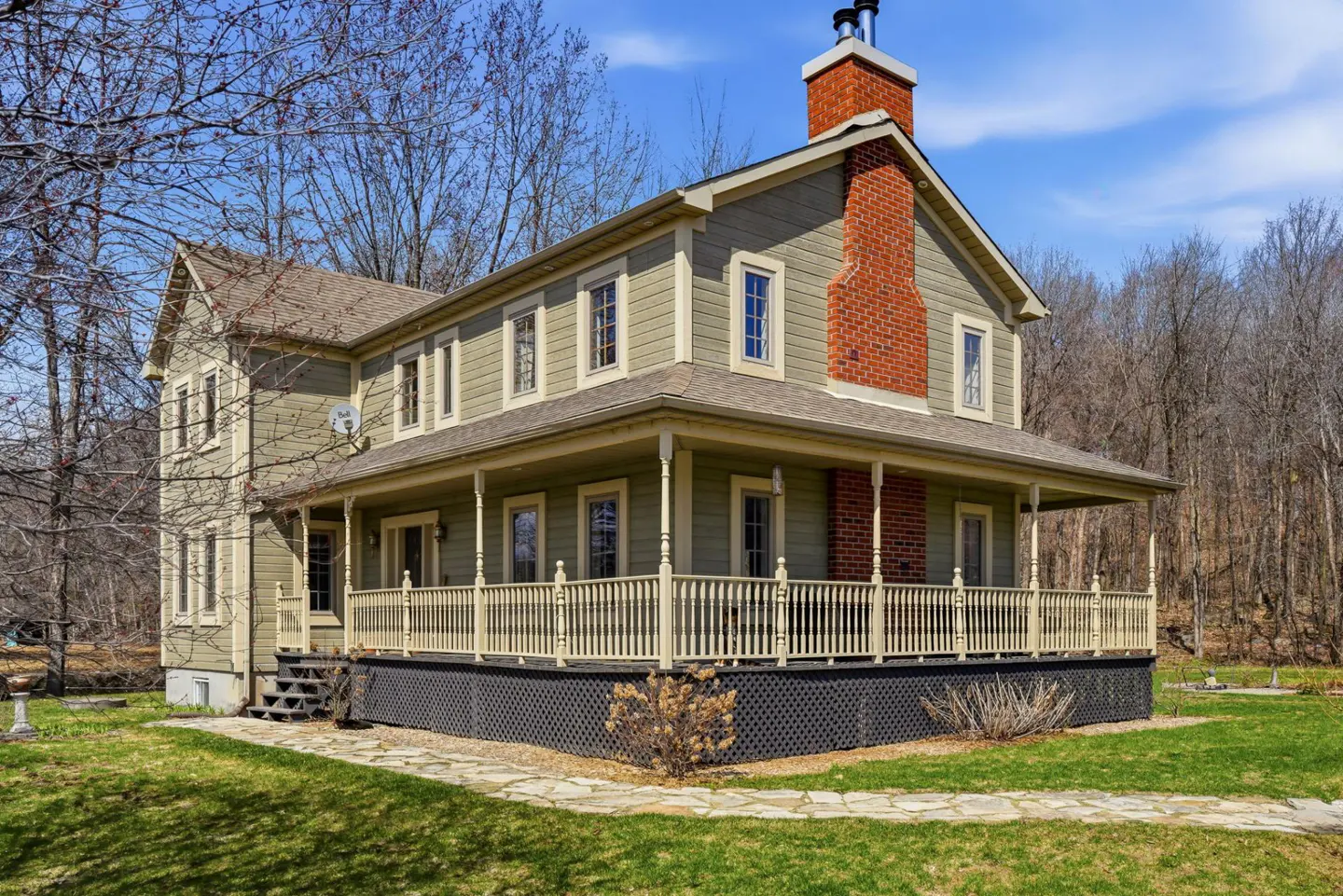 Two-story olive-green house with a brick chimney and a wraparound porch. The house is surrounded by a green lawn and trees.