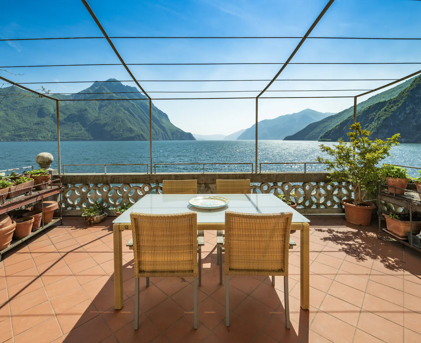 Outdoor patio with table and chairs overlooking a blue lake and mountains under a clear blue sky.