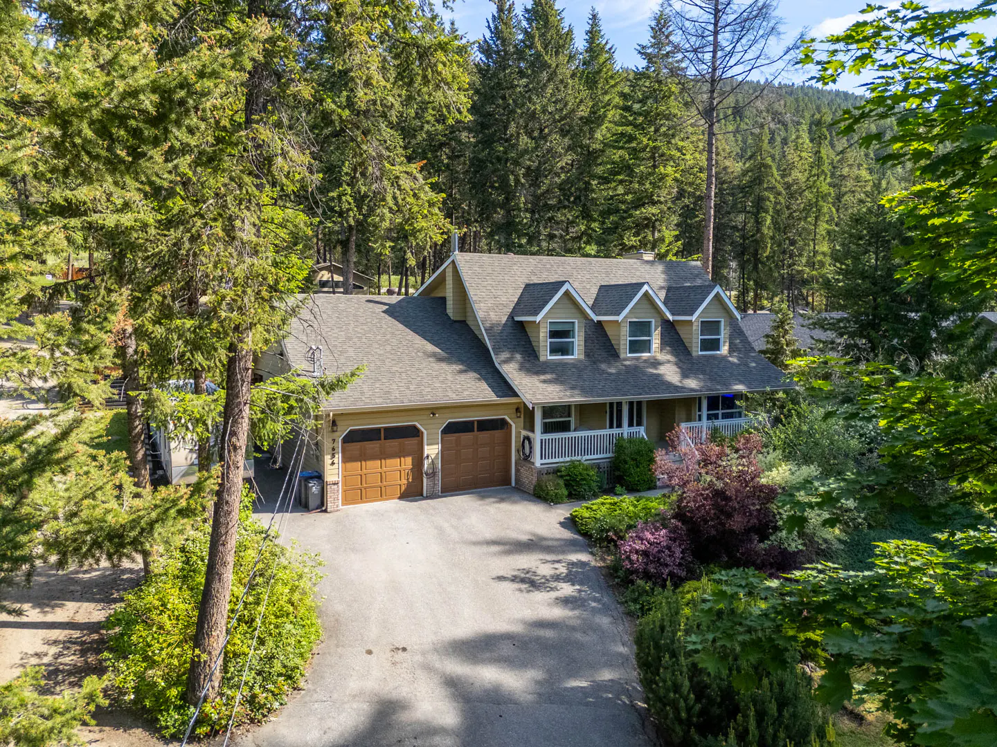 A yellow two-story house with a double garage and three dormer windows, nestled in a lush, pine-forested mountain setting.