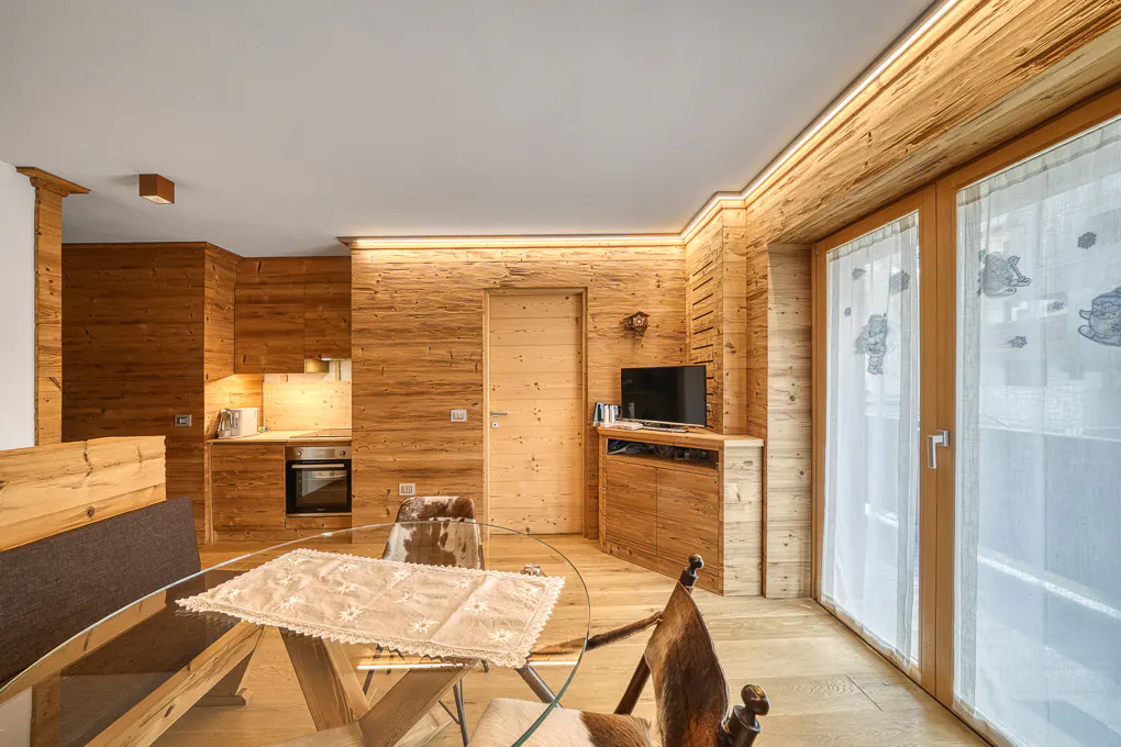 Cozy wood-paneled apartment with a glass-top table, cowhide chairs, and a small kitchen area. Natural light filters through sheer curtains.