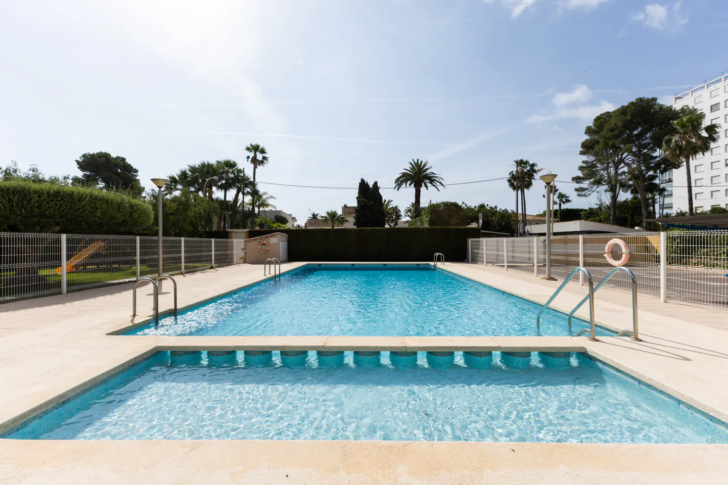 Outdoor pool with clear blue water, surrounded by a beige concrete deck, metal fence, palm trees, and a building in the background.