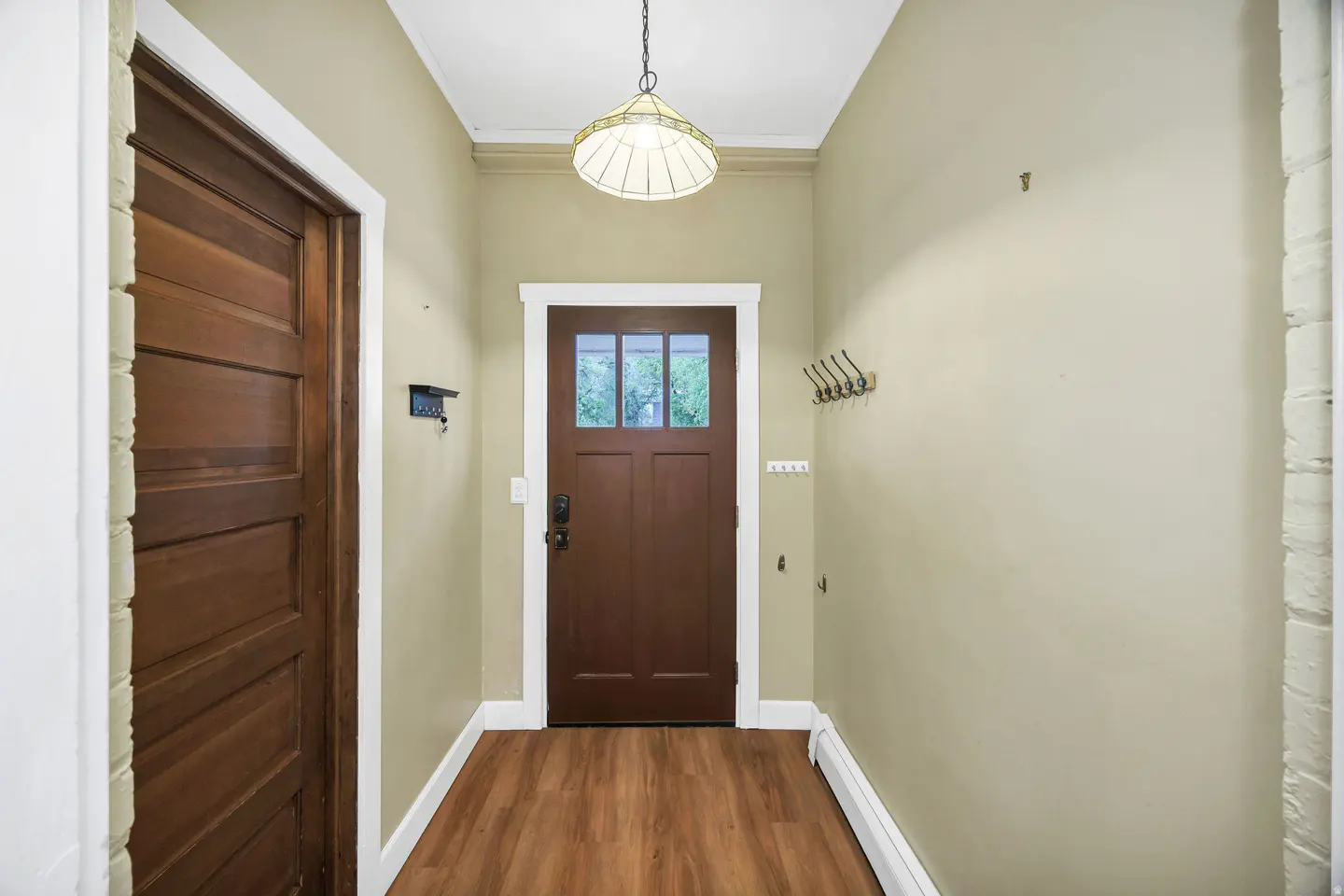 Entryway with brown wood floors, tan walls, and white trim. A brown front door with glass panes is centered. Coat hooks are on the right wall.