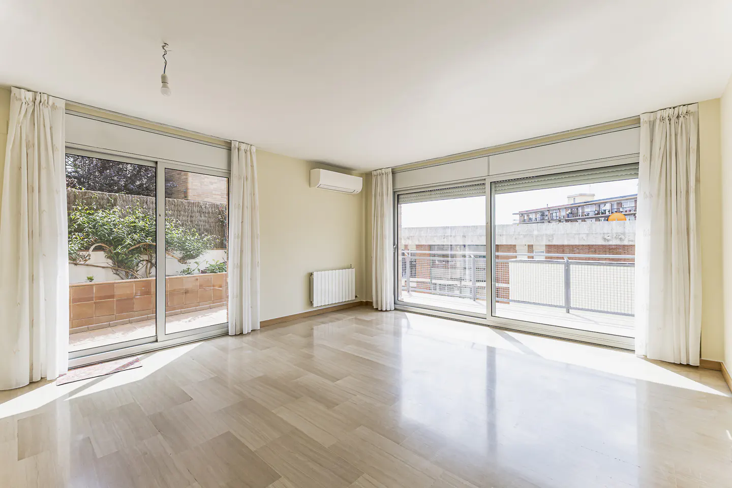 Bright, empty room with light wood floors, white walls, and large sliding glass doors with white curtains. Balcony and garden views.