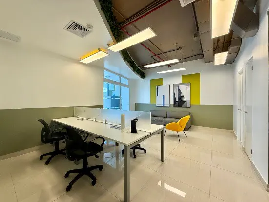 Bright office space with white desks, black chairs, and a gray sofa. Yellow accents include a chair and wall art. Exposed ceiling with ductwork.
