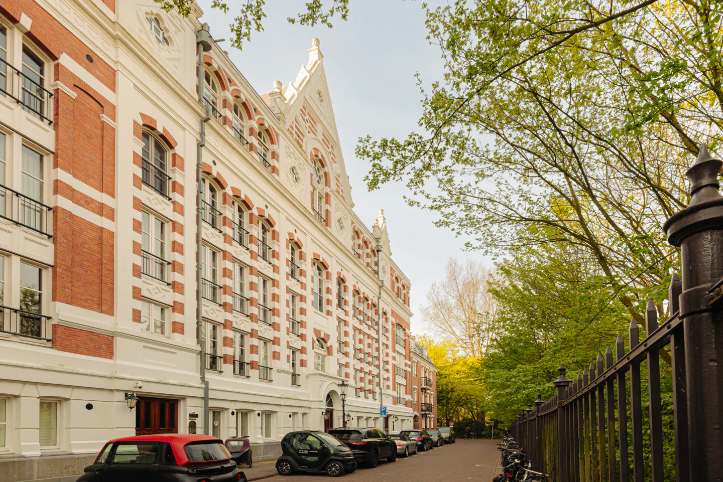 Exterior view of a red brick and white building with cars parked along the street. Trees are visible on the right.