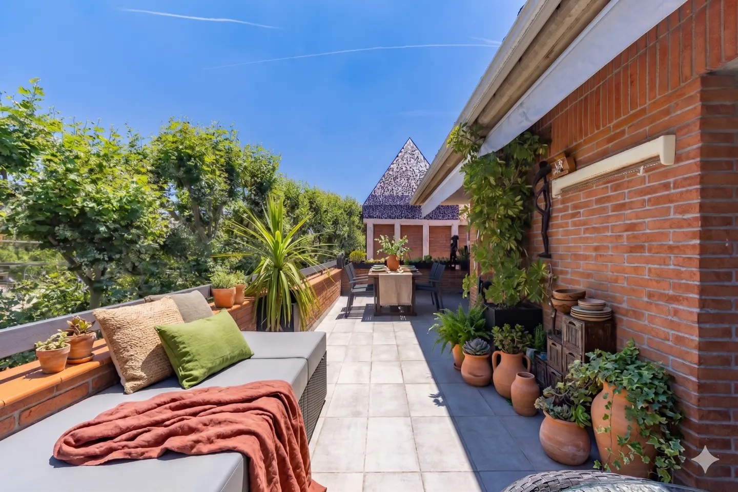 Sunny rooftop terrace with a grey sofa, terracotta pots, lush plants, and a dining table against a red brick wall.