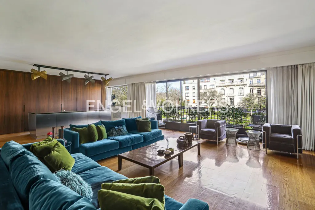 Bright living room with a blue sectional sofa, green pillows, wood floors, and a large window with a city view.