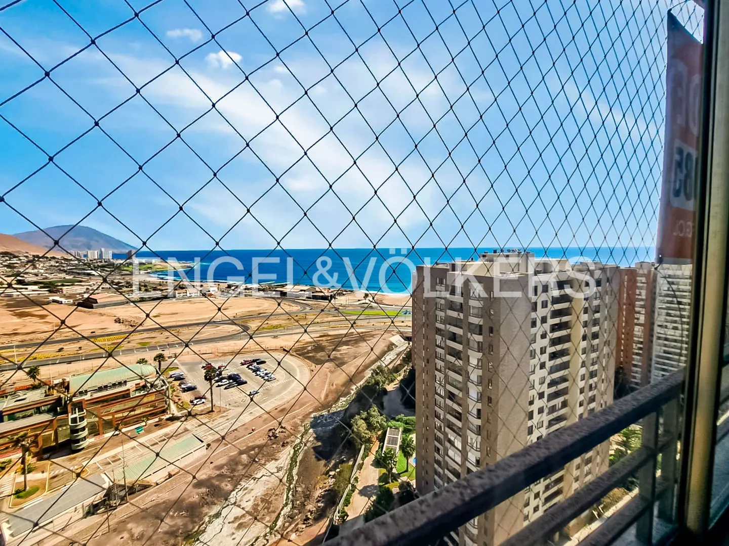 View from a high-rise balcony with safety netting, overlooking a coastal city, ocean, and mountains under a blue sky.