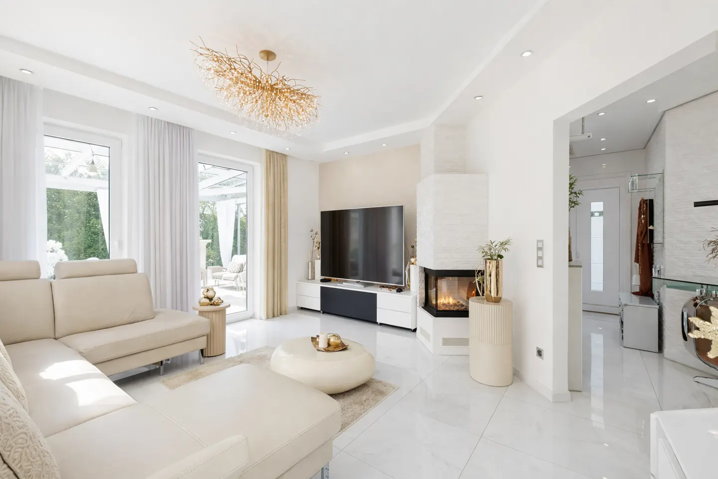 Bright, modern living room with a beige sectional sofa, white marble floors, and a fireplace. A large TV sits above a console table. A gold chandelier hangs from the ceiling.