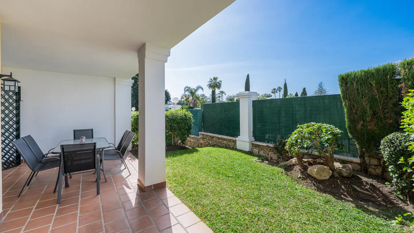 Covered patio with a table and chairs overlooks a green lawn and garden under a blue sky.