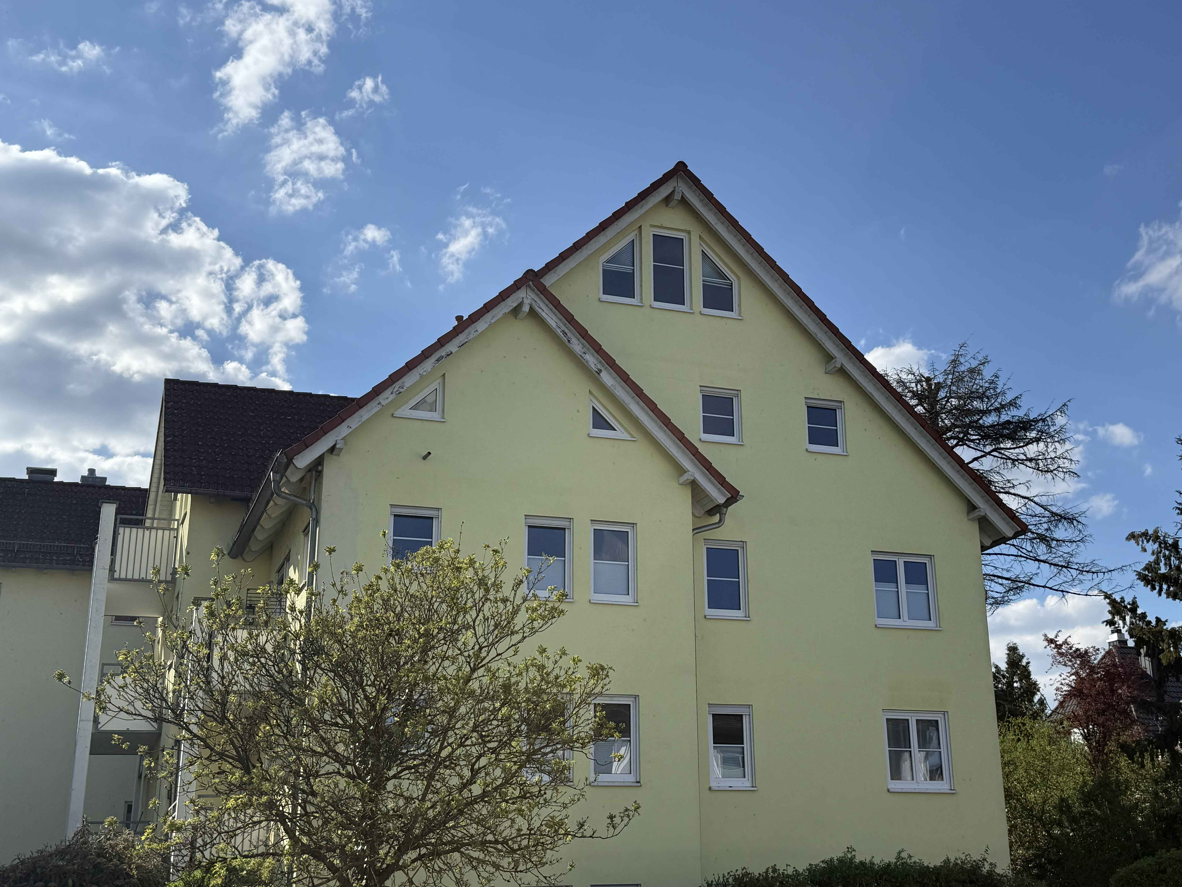 A pale yellow apartment building with a brown roof and white window frames under a blue sky with fluffy white clouds.