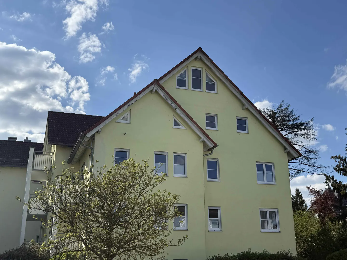 A pale yellow apartment building with a brown roof and white window frames under a blue sky with fluffy white clouds.
