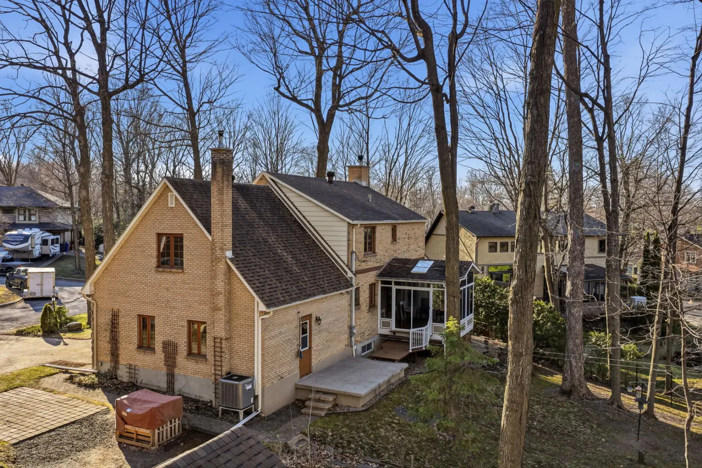 Back of a tan brick house with a brown roof, surrounded by trees and other houses on a sunny day.