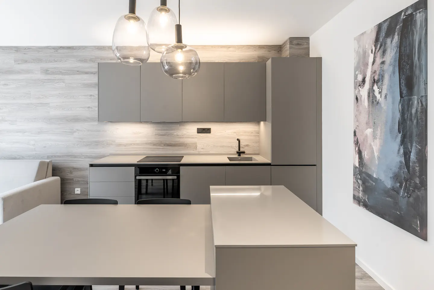 Modern kitchen with gray cabinets, island, and dining table. Three pendant lights hang above the island. An abstract painting adorns the white wall.