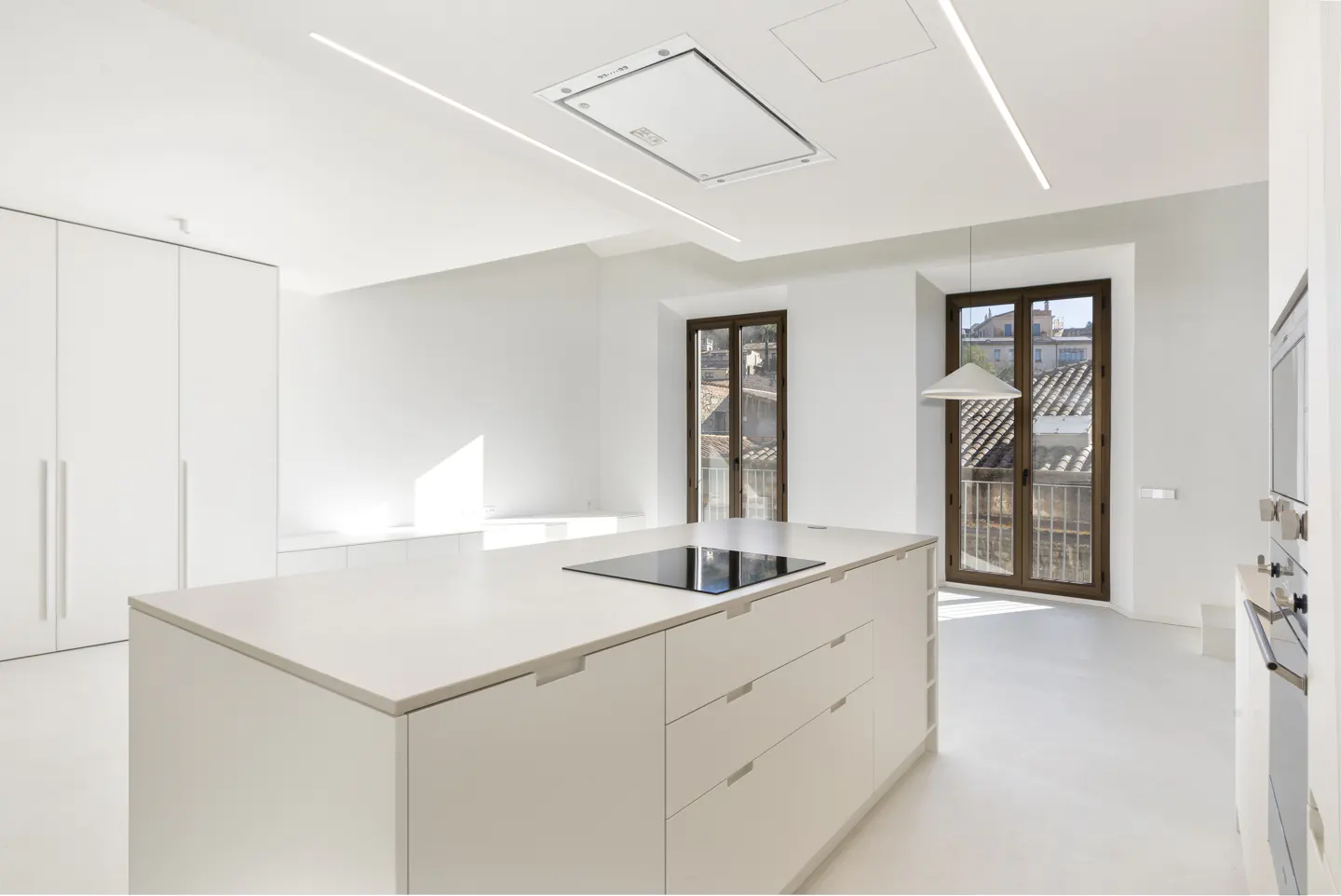 Bright, all-white kitchen with a large island, cooktop, and two windows with brown frames overlooking a cityscape.
