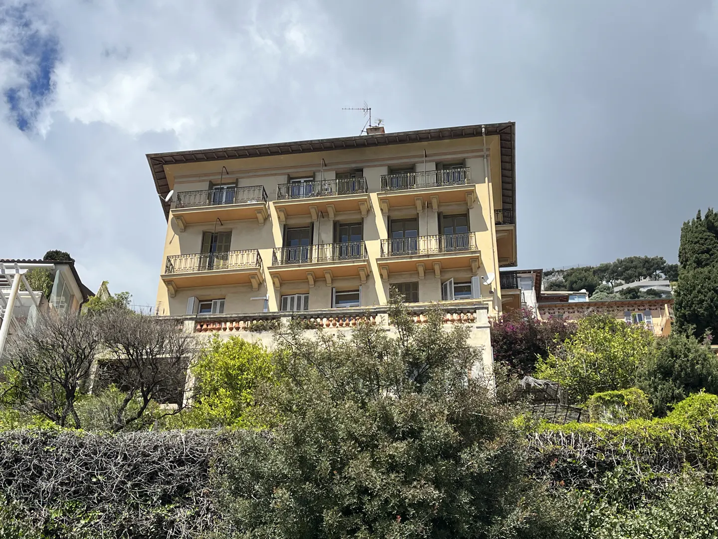A low-angle shot of a tan building with black balconies, surrounded by green trees and bushes.