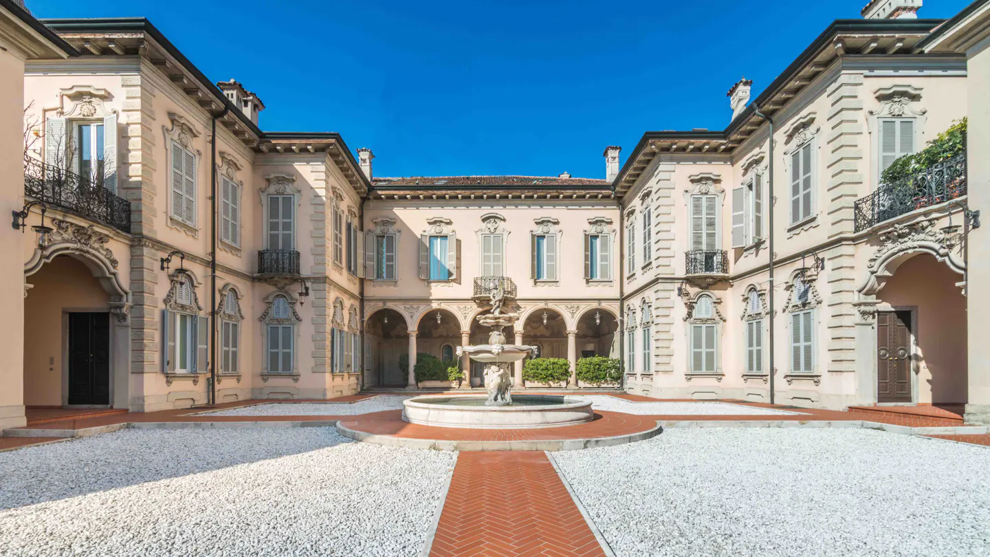 Courtyard view of a pale pink building with white shutters and a fountain in the center. The ground is covered in white gravel.