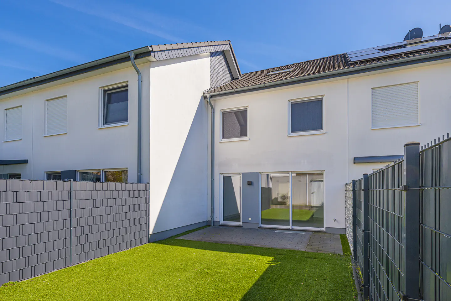 Backyard view of a modern, white townhouse with a small patio and green lawn, enclosed by gray fences under a clear blue sky.