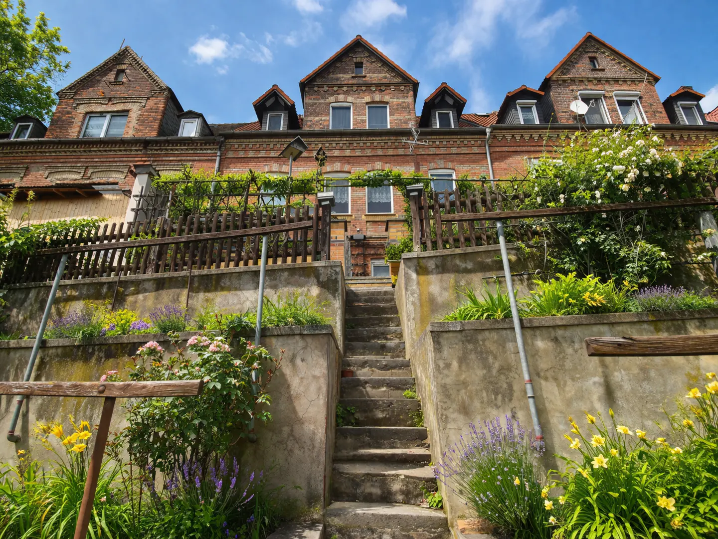 Three brick houses sit above a concrete retaining wall and stairs, with flowers and greenery in the foreground.