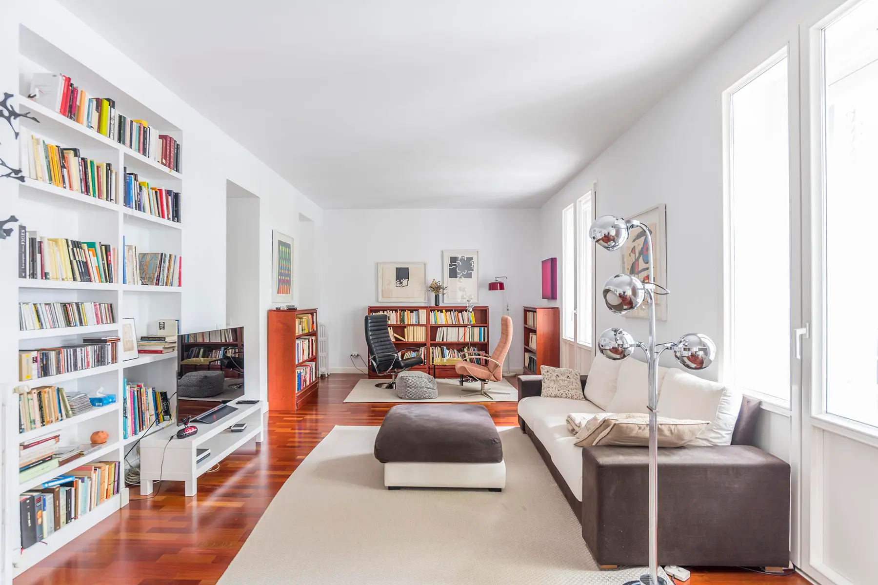 Bright living room with white walls, hardwood floors, a white sofa, a large bookshelf, and a modern chrome floor lamp. [AI-generated text]