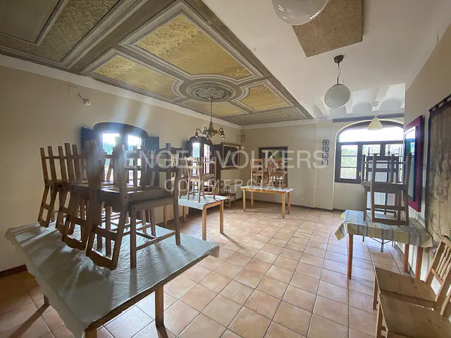 A spacious room with chairs stacked on tables, terracotta tile floor, and decorative ceiling. Natural light streams through arched windows.