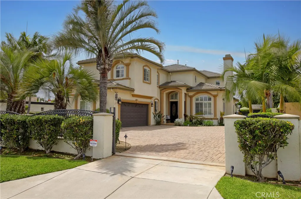 Beige two-story house with brown trim, a brown garage door, and a brick driveway. Palm trees and green bushes surround the house.