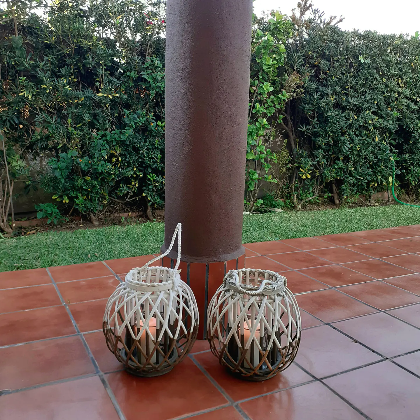 Two round, wicker lanterns with candles inside sit on a red-tiled patio near a brown pillar and green bushes.