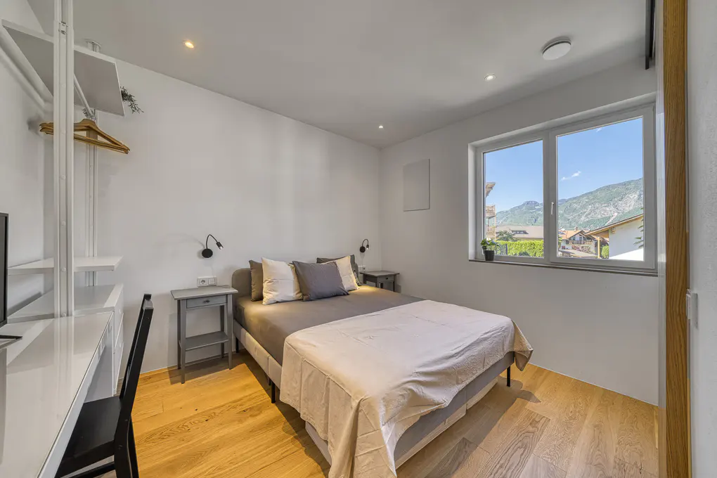 Bright bedroom with a gray bed, white walls, and wood floors. A desk and chair are on the left, and a window with a mountain view is on the right.