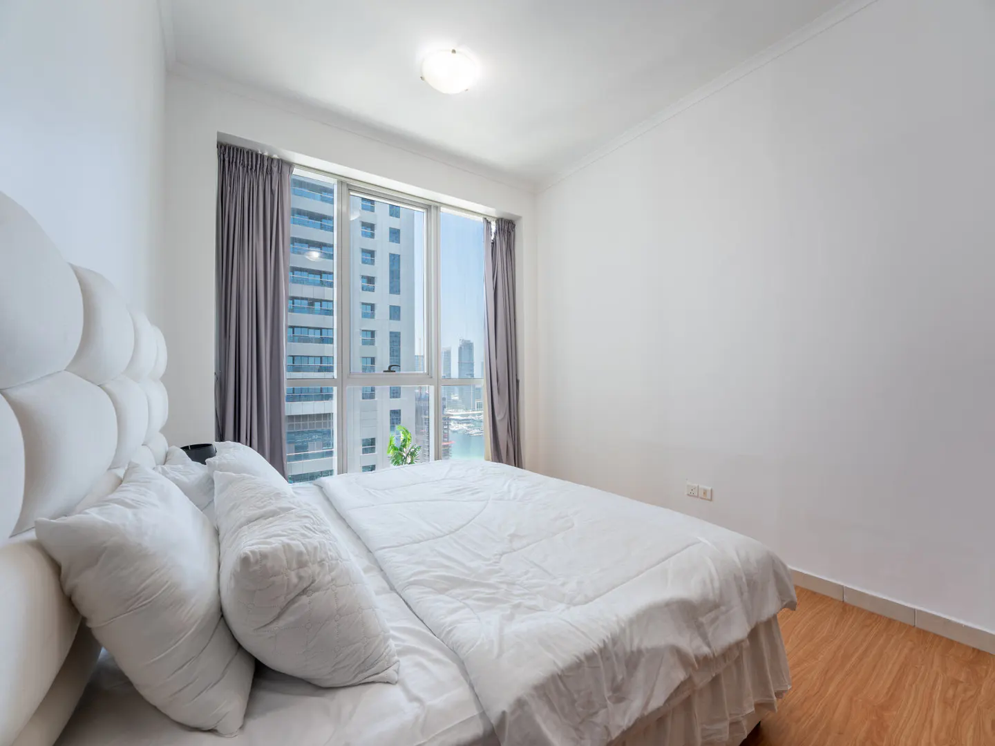 Bright bedroom with white walls, wood floor, and a large window with city views. The bed has a white headboard and bedding. Gray curtains frame the window.
