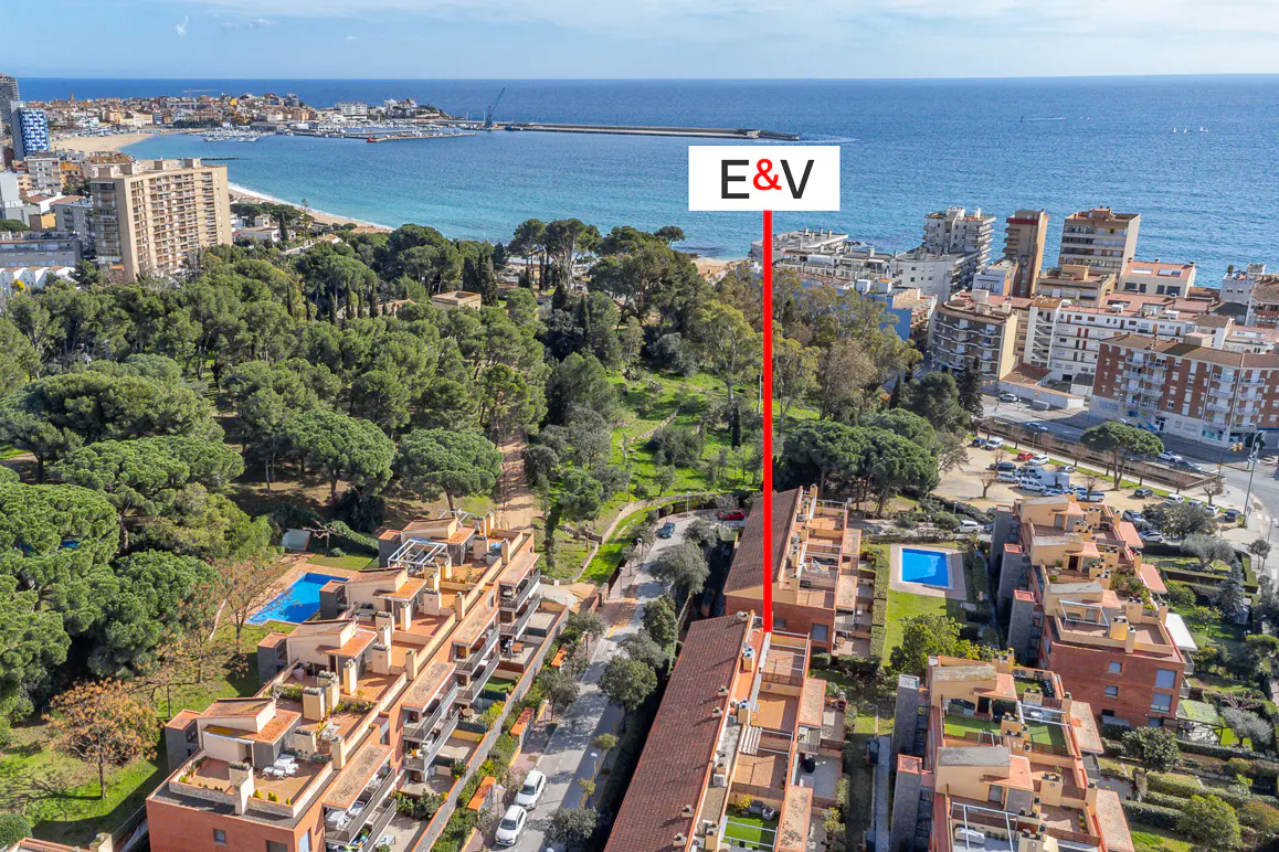 Aerial view of a coastal town with apartment buildings, trees, and the sea in the background. A red line points to a building with "E&V" above it.