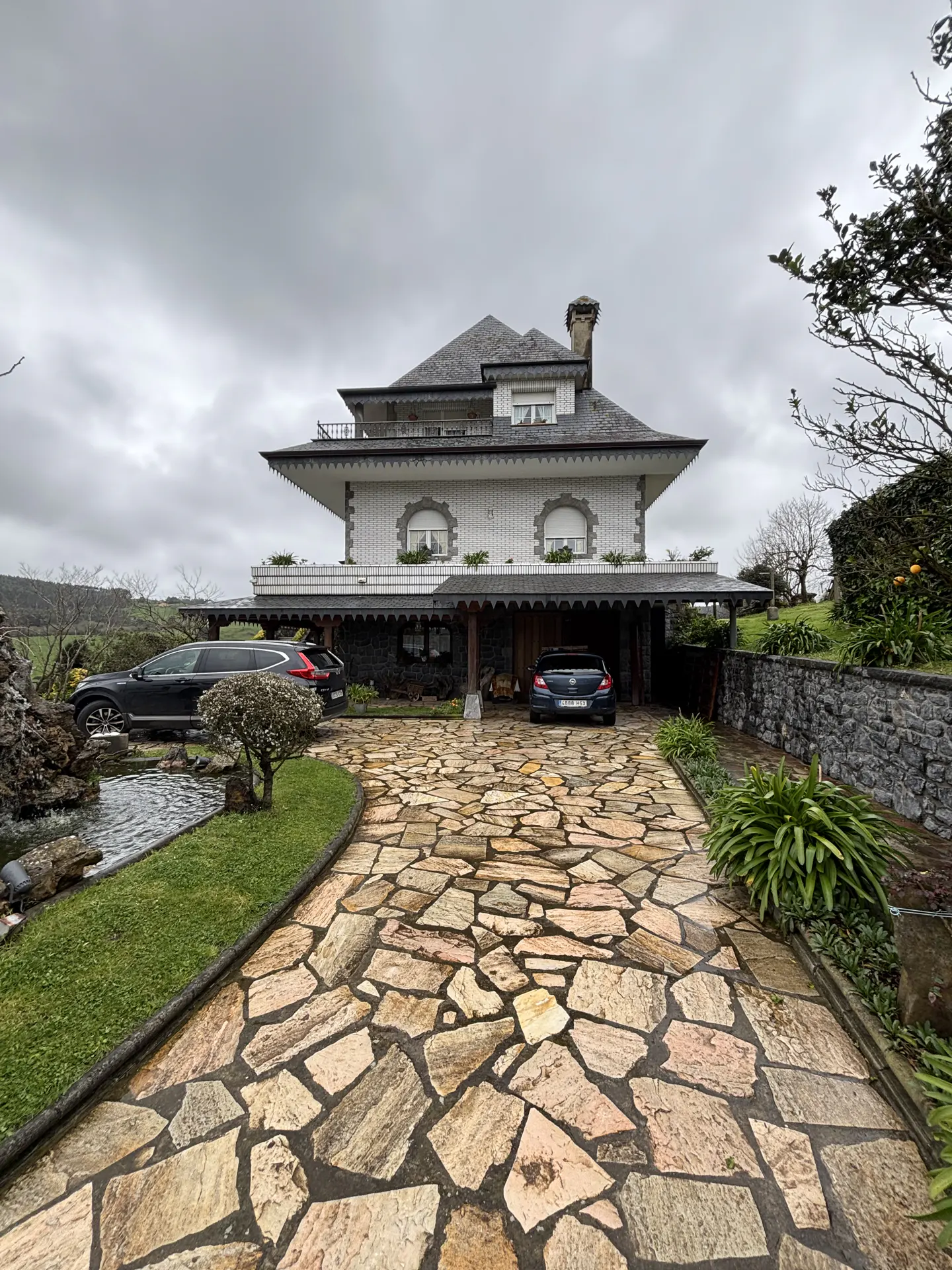 Two-story white house with a gray roof and stone driveway. Two cars are parked in front of the house. Cloudy sky.