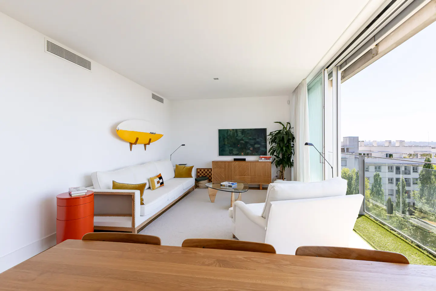 Bright living room with white walls, sofa, chair, and a yellow surfboard decoration. Balcony with city view.