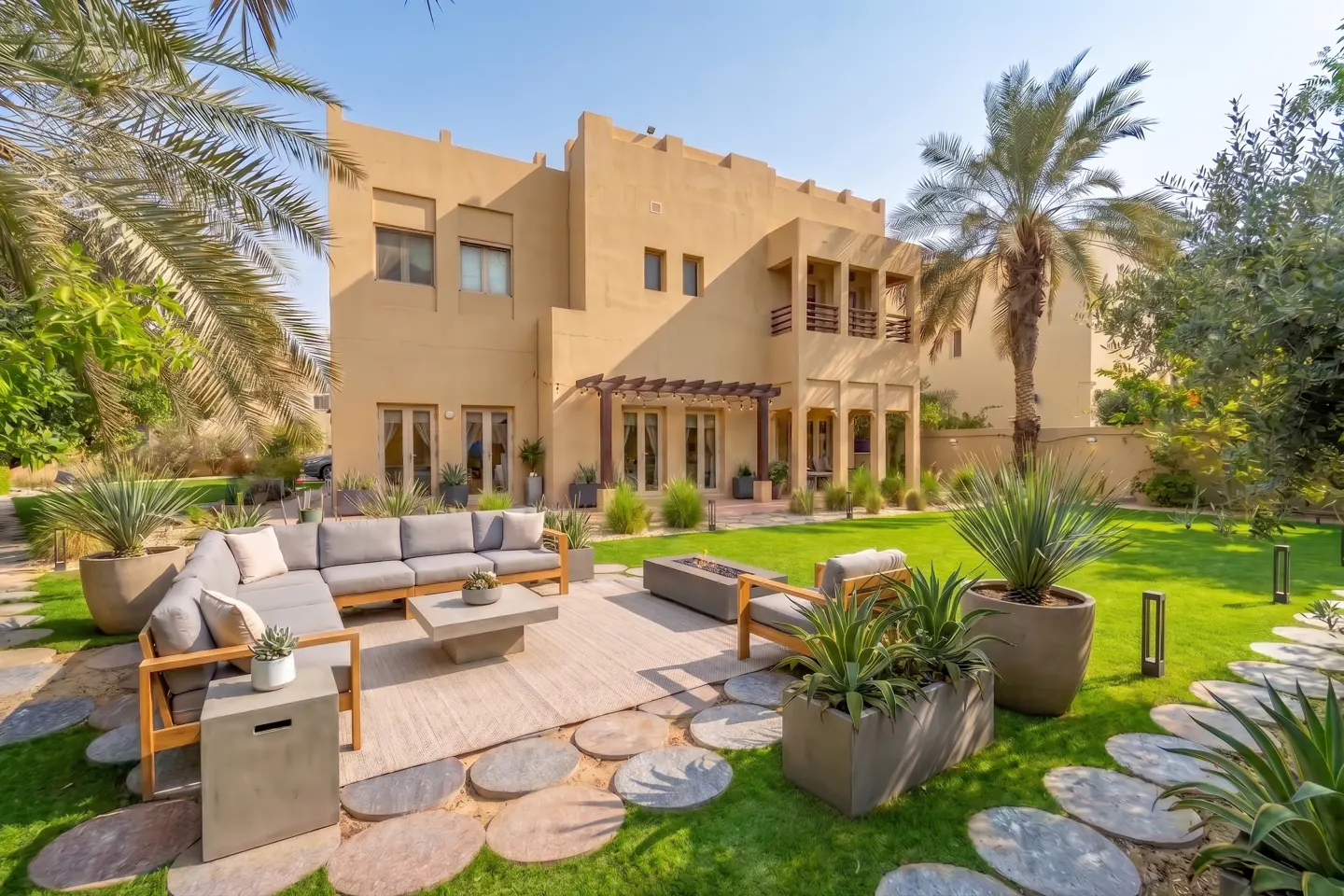 Backyard patio with a tan villa, sectional sofa, fire pit, and lush green lawn with stone pavers and palm trees.
