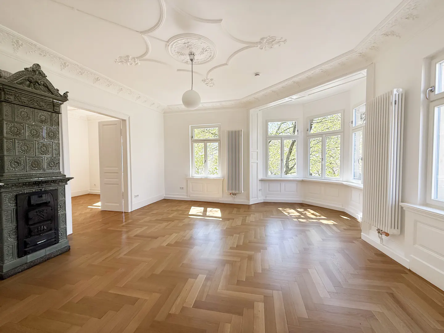 Bright, empty room with herringbone wood floors, white walls, and a tiled fireplace. A bay window offers a view of trees.