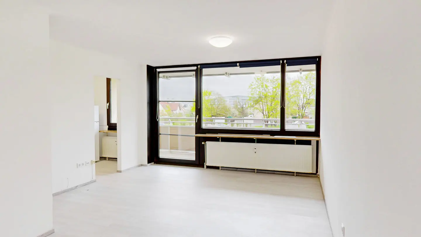 Bright, empty room with white walls, light gray floor, and black-framed windows overlooking a balcony and trees.