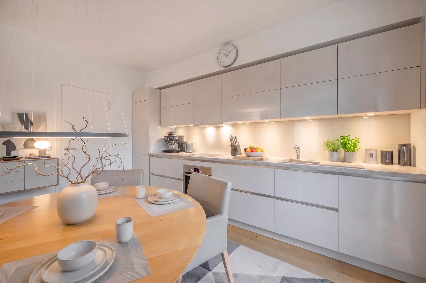 Modern kitchen with light gray cabinets, wooden dining table, and minimalist decor under warm under-cabinet lighting.