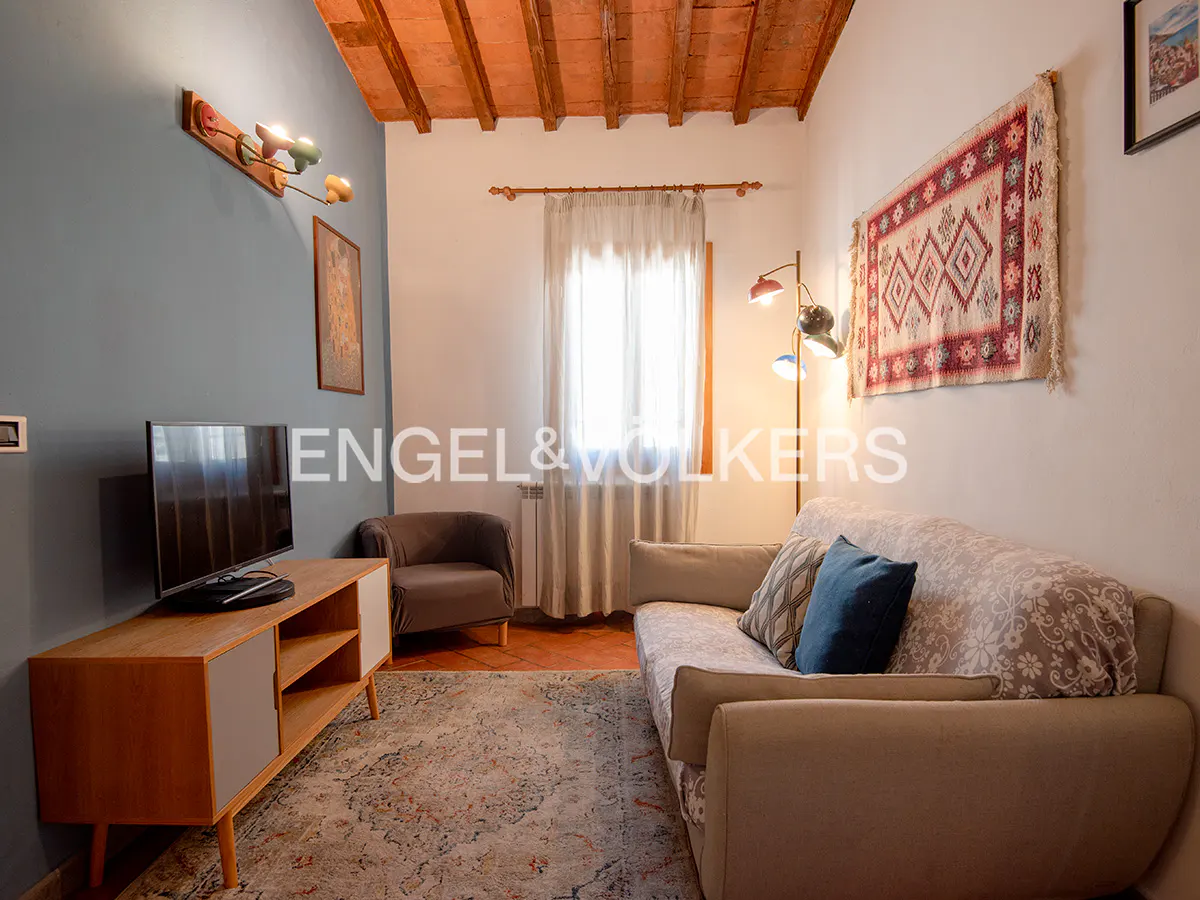Living room with a beige sofa, patterned rug, TV on a wooden stand, and a brown armchair. A colorful tapestry hangs on the wall.