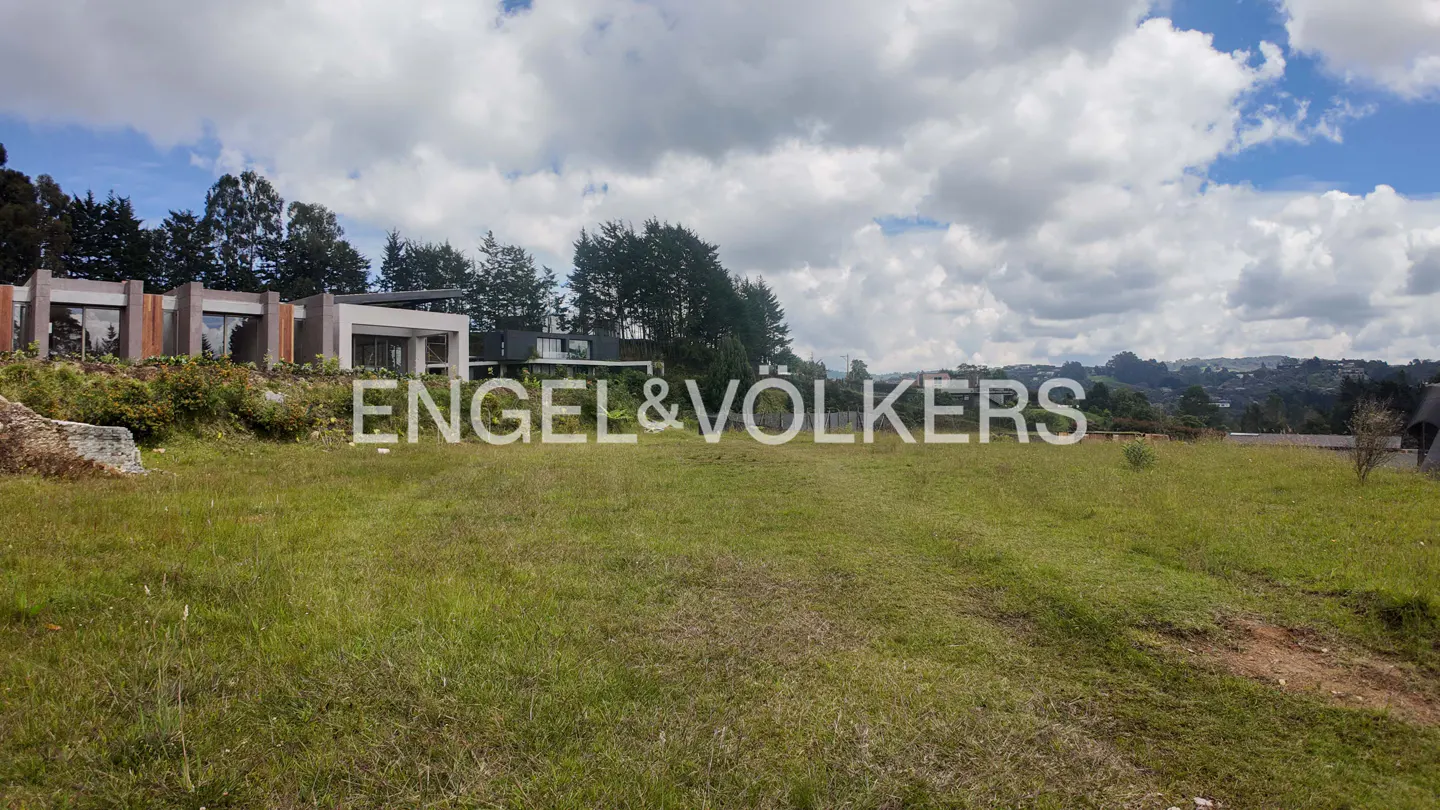 Lush green field with modern homes in the background under a cloudy sky. Engel & Volkers logo overlay.