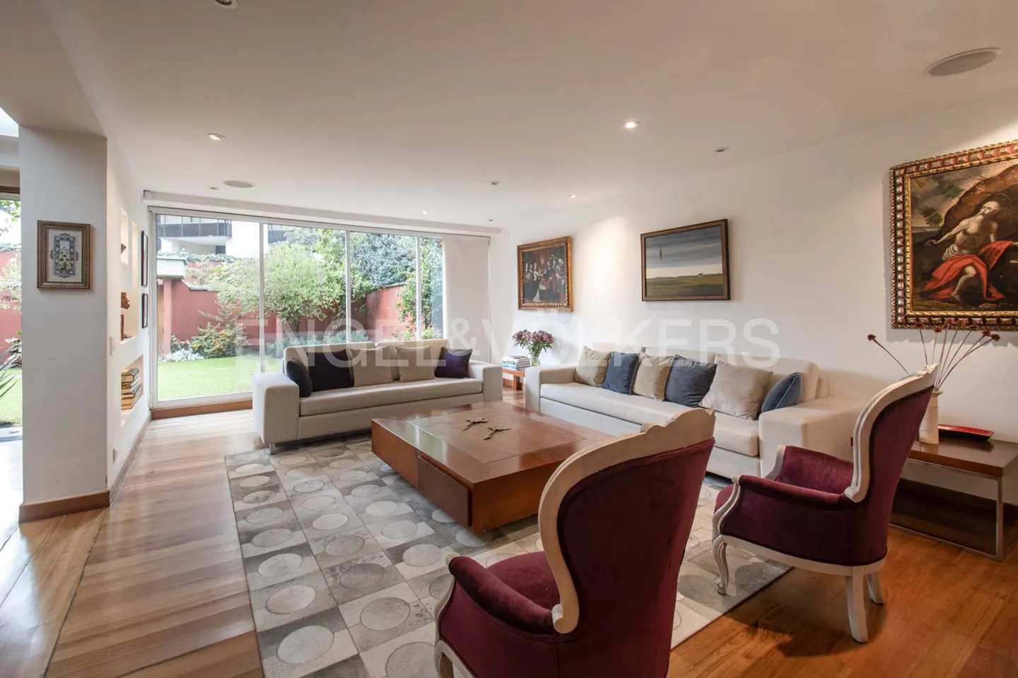 Bright living room with two white sofas, a wood coffee table, and two burgundy chairs on a patterned rug. Large windows overlook a green garden.
