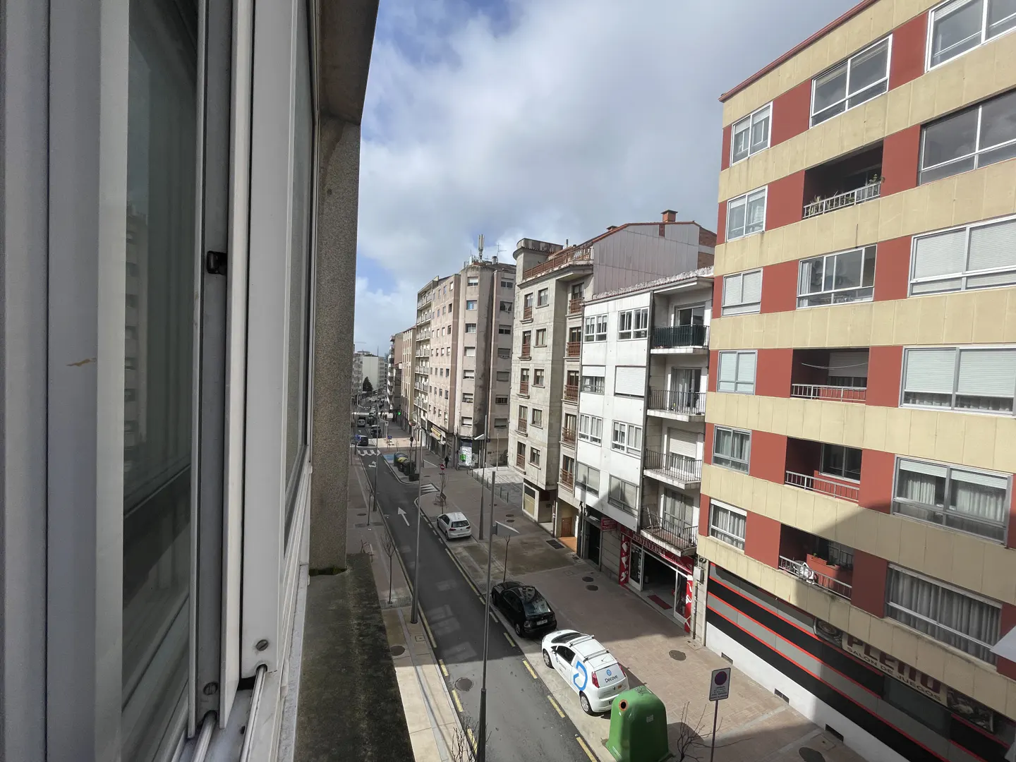View from a window overlooking a city street with cars, buildings, and a cloudy sky.