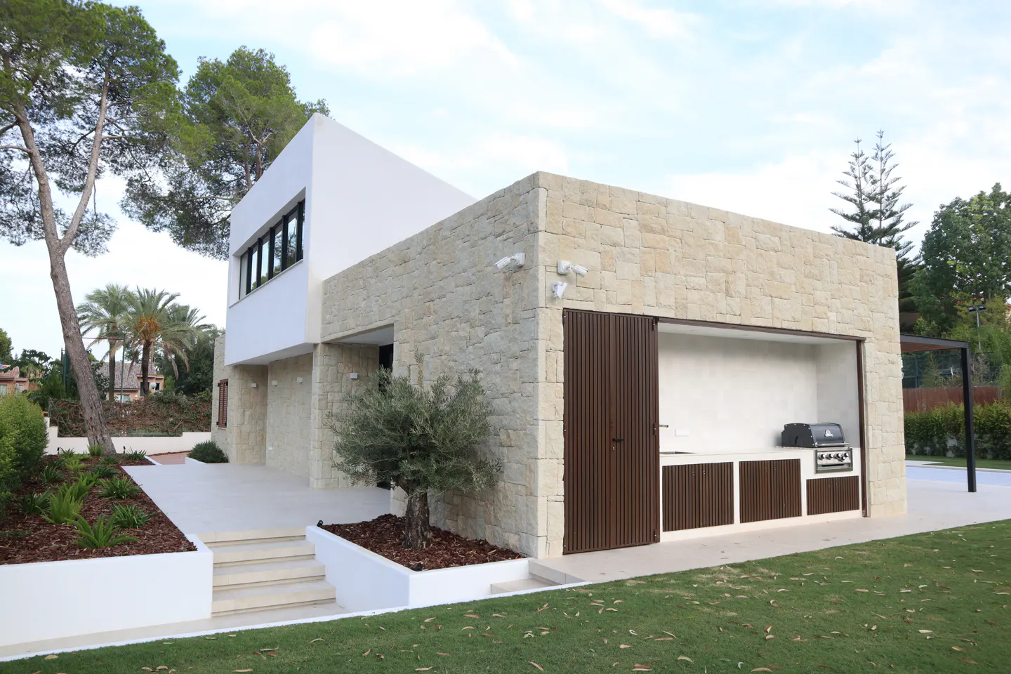 Modern home exterior with a stone facade, white walls, and a built-in outdoor kitchen with a grill. Green lawn and trees surround the property.