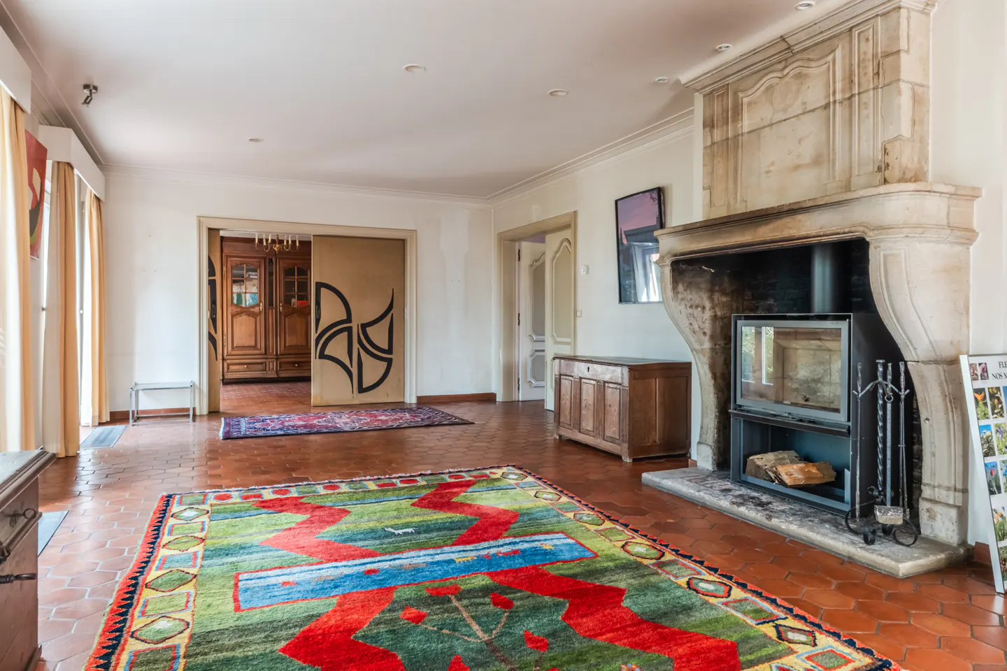 A spacious living room with a stone fireplace, colorful rug, and terracotta tile flooring.