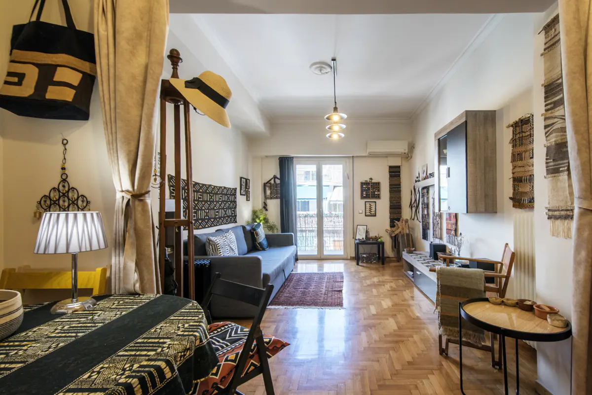 Interior view of a living room with a gray sofa, wooden floors, and ethnic-inspired decor. A table with a patterned tablecloth is in the foreground.