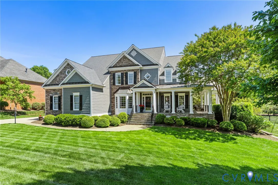 Two-story house with gray siding, stone accents, black shutters, and a covered front porch with white columns. Manicured lawn and blue sky.