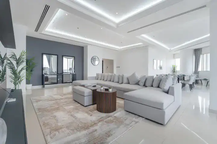 Bright, modern living room with a large gray sectional sofa, round wood table, and beige rug. Dining area with white chairs visible in background.